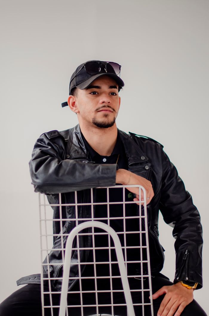 featured-images-02 A confident man posing with a chair in a leather jacket and cap, exuding modern style.