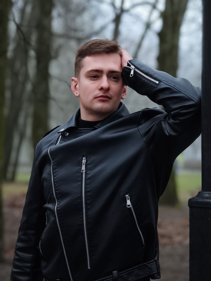 why-choose-us-02 Young man in a leather jacket posing by a lamppost in an autumn park setting.