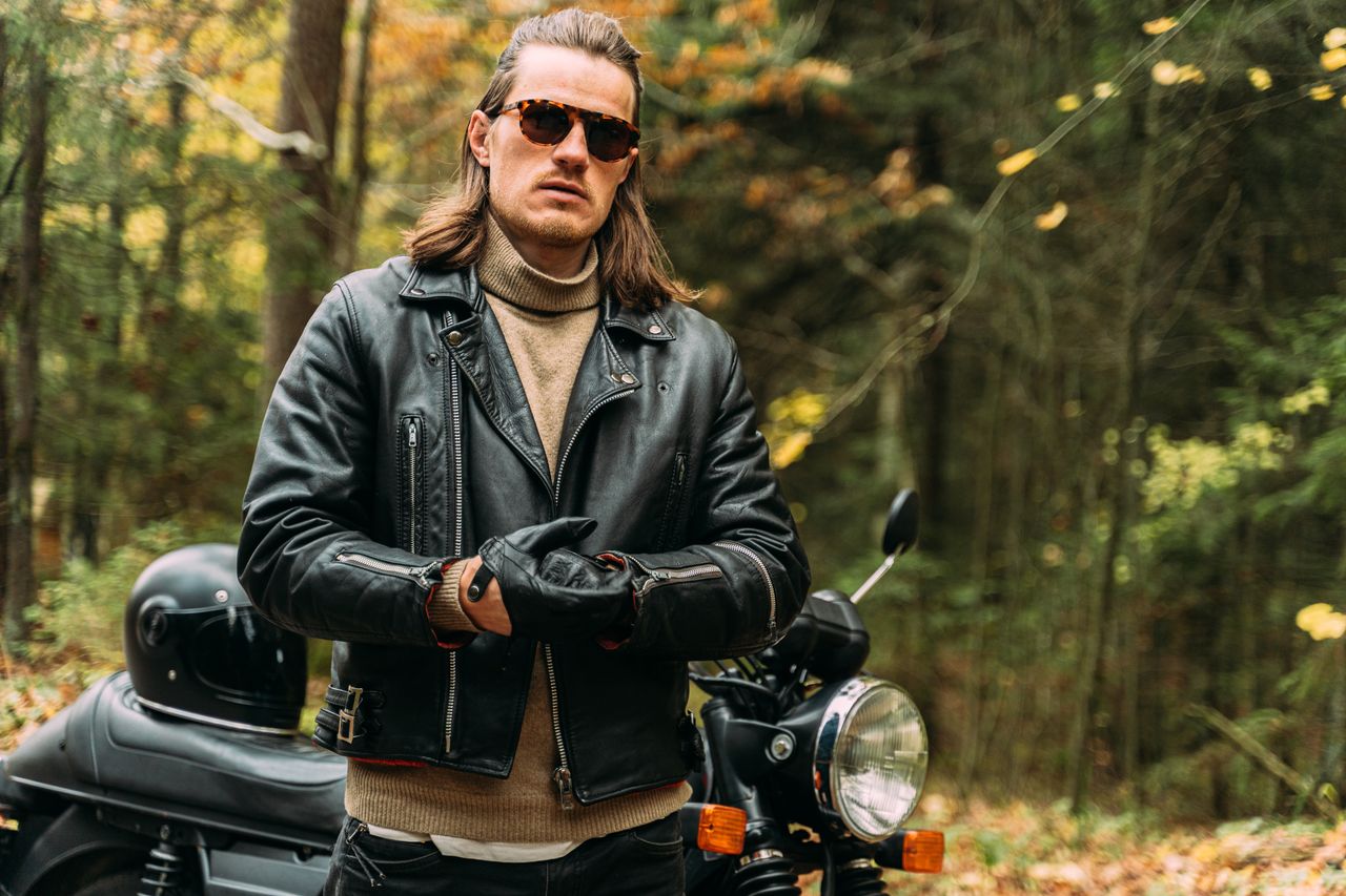 gallery-02 Confident biker in black leather jacket standing by motorcycle in a forest setting.