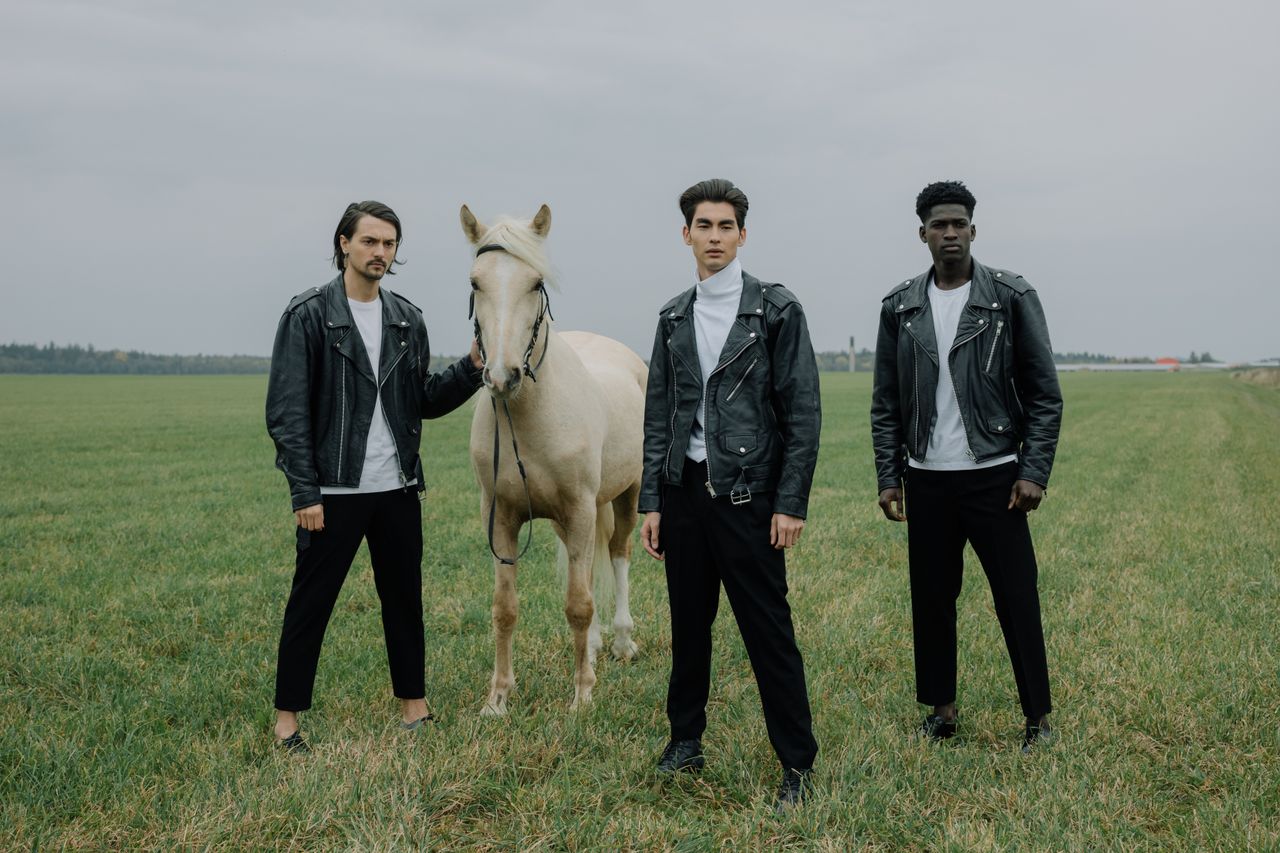 services-01 Three men in black leather jackets pose with a horse on a grass field.