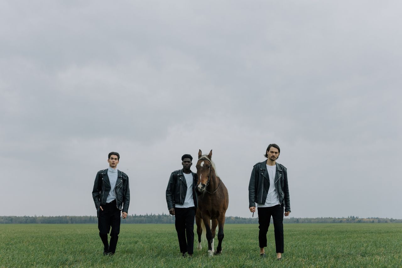gallery-07 Three diverse men in leather jackets with a horse in an open field under a cloudy sky.