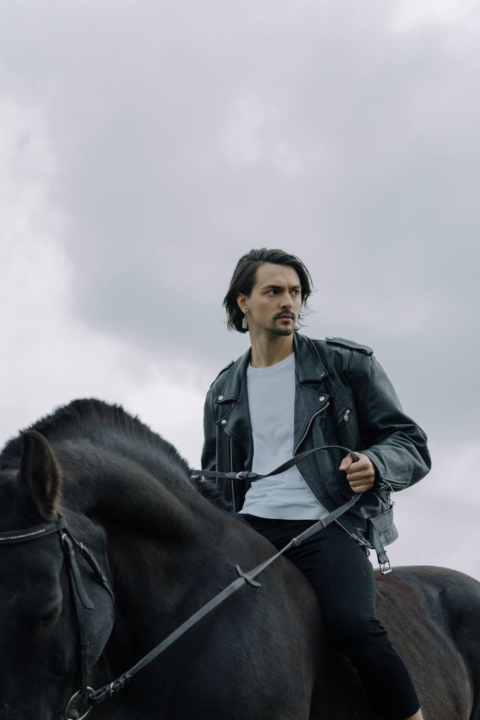 services-03 A man in a black leather jacket riding a horse under a cloudy sky. Dramatic and adventurous vibe.