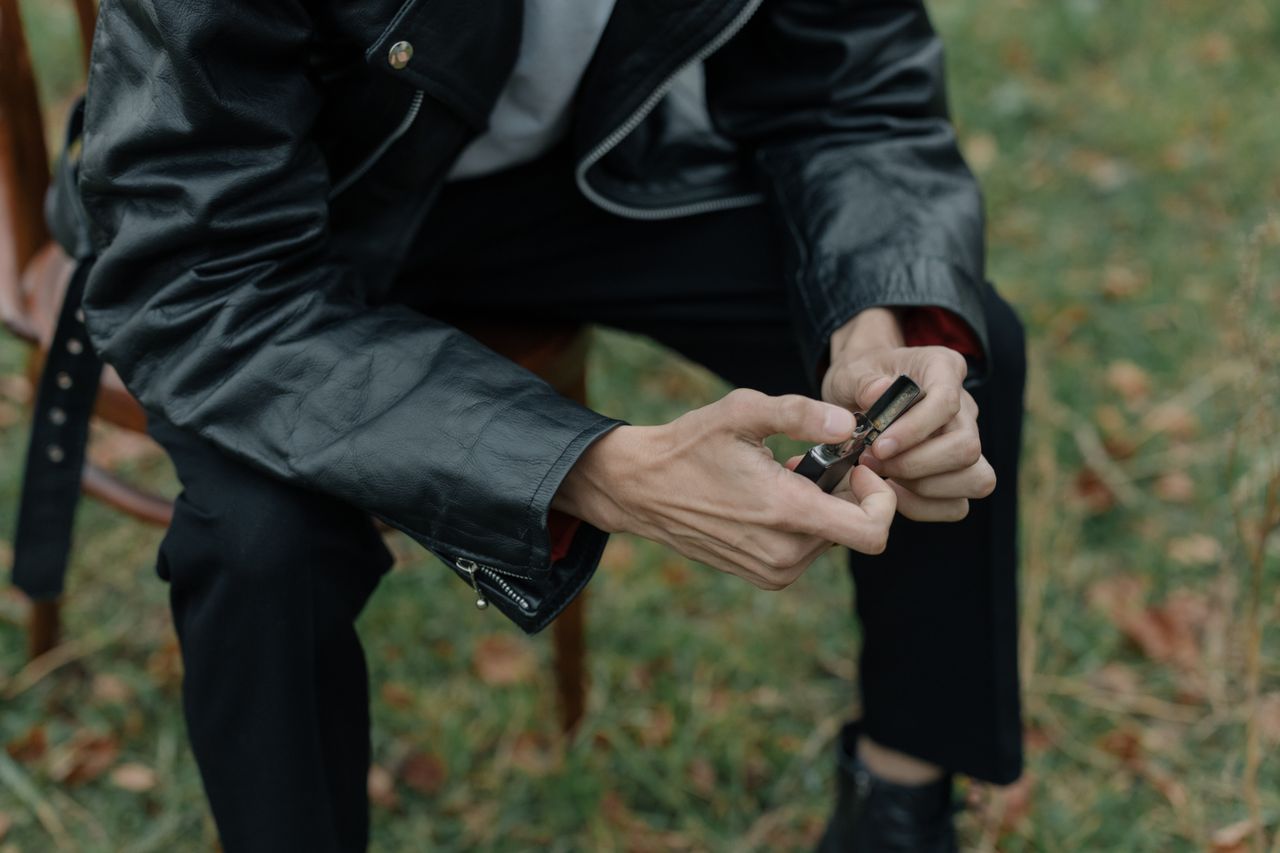 about-me-01 A person in a leather jacket holding a lighter while sitting outdoors on grass.